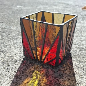 May include: A square stained glass candle holder with a geometric design. The glass panels are in shades of red, orange, and yellow, with black lines separating the pieces. The candle holder casts a colorful shadow on the surface.