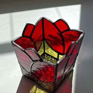 May include: A red stained glass candle holder shaped like a flower. The holder has a yellow center and is made of small pieces of glass.