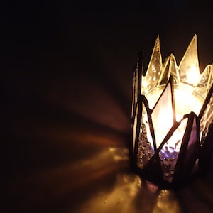 May include: A glass candle holder shaped like a crown with a lit candle inside. The candle holder is made of stained glass with a purple and clear design.