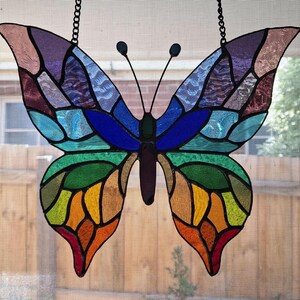 May include: A stained glass butterfly with a rainbow color scheme. The butterfly is made of many small pieces of glass, each with a different color. The butterfly is hanging from a chain.
