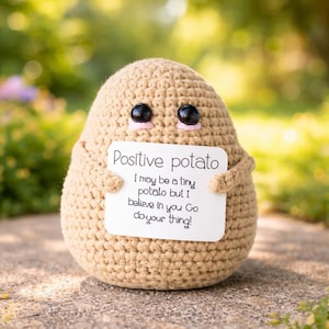Puede incluir: Una figura de patata tejida a ganchillo con ojos negros y un pequeño rubor rosa. Sostiene un letrero que dice "Positive potato" y un mensaje motivador. La figura es de color beige y está sobre una superficie de piedra.