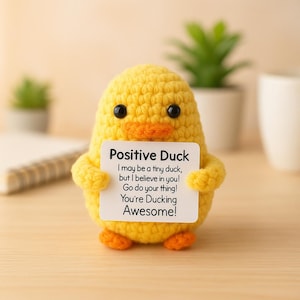 May include: A handmade yellow crochet duck with black eyes and an orange beak and feet. The duck holds a white sign with the text "Positive Duck" and a motivational message. A charming, whimsical item.