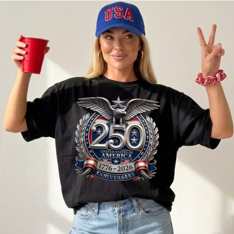 Pode incluir: Camiseta preta com um design patri&oacute;tico que comemora o 250&ordm; anivers&aacute;rio dos Estados Unidos da Am&eacute;rica. O design inclui uma &aacute;guia, estrelas e o texto "250 United States of America 1776-2026 Anniversary". Um bon&eacute; de beisebol azul "USA" tamb&eacute;m &eacute; vis&iacute;vel.