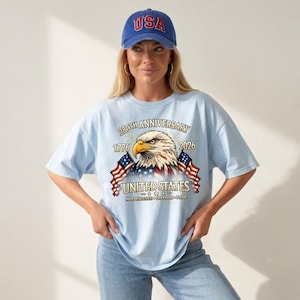 May include: A light blue t-shirt with a graphic of an eagle, American flags, and the text "250th Anniversary 1776-2026 United States Independence Freedom Pride." The person is wearing a blue baseball cap with "USA" on it.