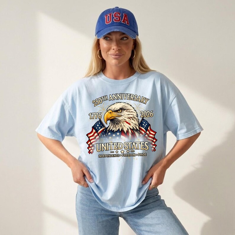 May include: A light blue t-shirt with a graphic of an eagle, American flags, and the text "250th Anniversary 1776-2026 United States Independence Freedom Pride." The person is wearing a blue baseball cap with "USA" on it.