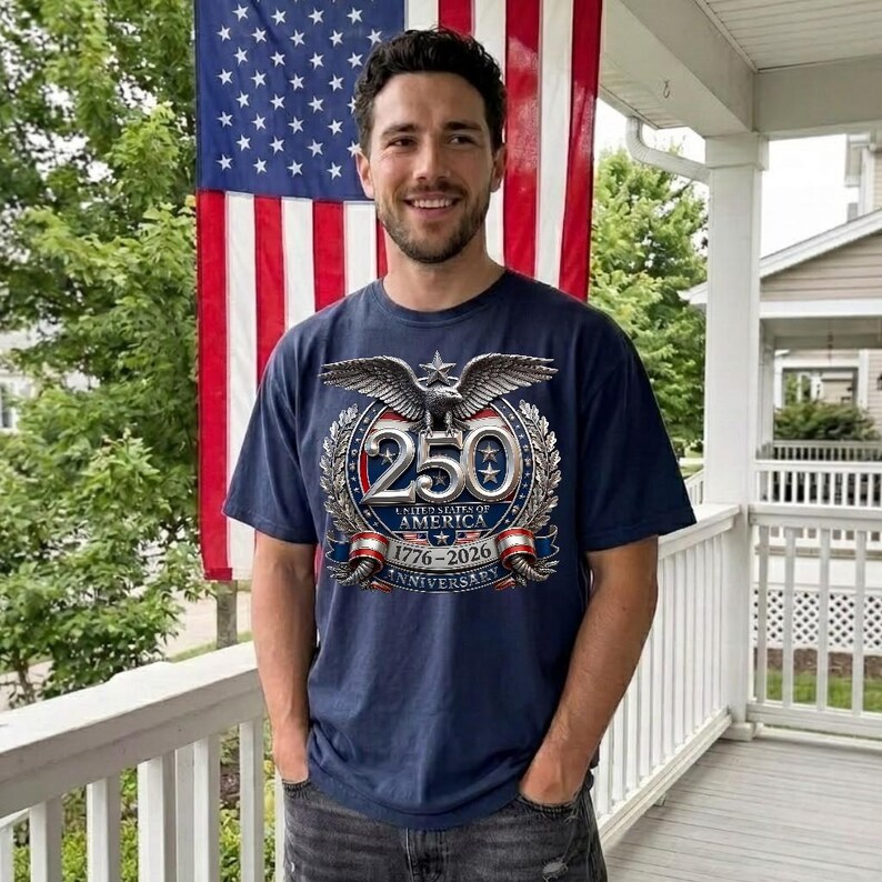 Pode incluir: Camiseta azul marinho com um emblema de &aacute;guia prateada e o texto "250 United States of America 1776-2026 Anniversary". A pessoa usa a camiseta em frente a uma bandeira americana.