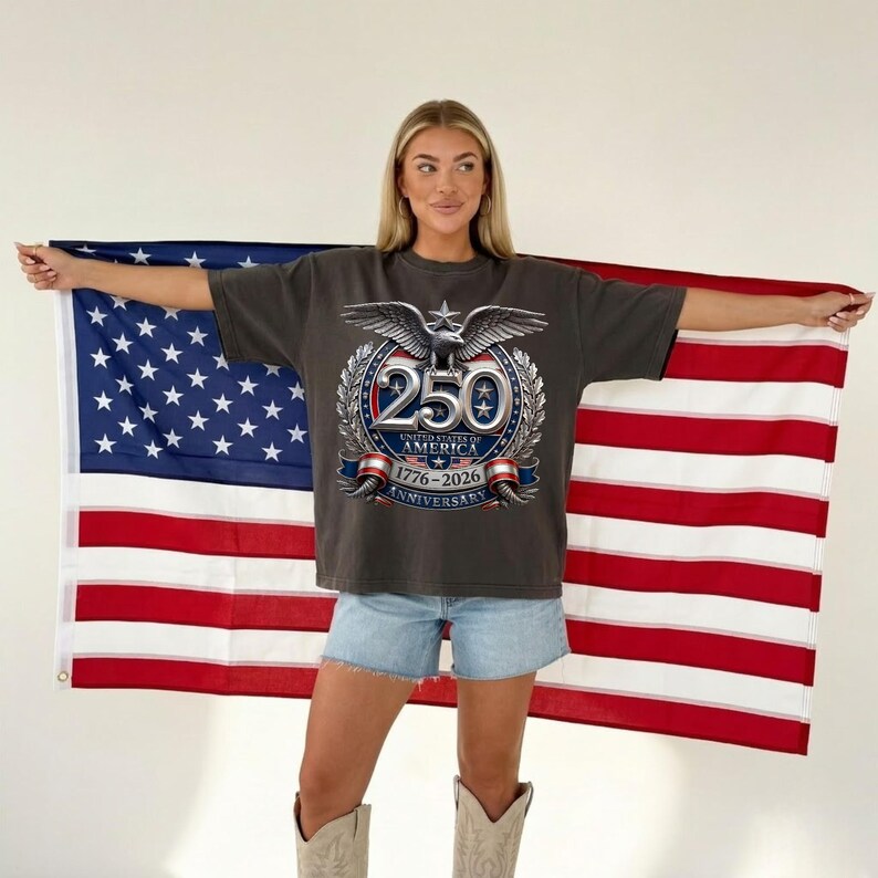 Pode incluir: T-shirt cinzenta escura com um gr&aacute;fico que comemora o 250&ordm; anivers&aacute;rio dos Estados Unidos da Am&eacute;rica. O design apresenta uma &aacute;guia, estrelas e os anos 1776-2026. A pessoa segura uma bandeira americana.