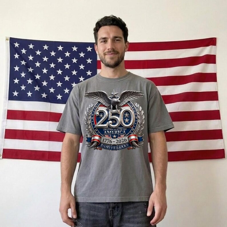 Pode incluir: Uma camiseta cinza com um gr&aacute;fico comemorativo do 250&ordm; anivers&aacute;rio dos Estados Unidos da Am&eacute;rica. O design apresenta uma &aacute;guia, o n&uacute;mero 250 e os anos 1776-2026. A bandeira americana est&aacute; ao fundo.