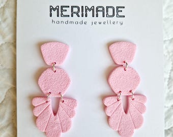 May include: A pair of pink polymer clay earrings with a geometric design. The earrings are made up of three different shapes: a small circle, a larger hexagon, and a fan-shaped piece. The earrings are hanging from silver hooks. The earrings are displayed on a white card with the text "MERIMADE handmade jewellery" and "@merimade_clay".
