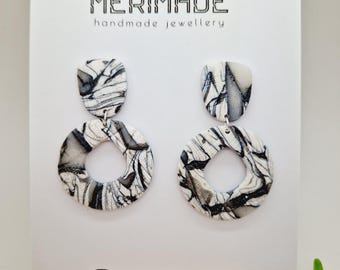 May include: A pair of black and white marbled dangle earrings with a round shape and a smaller top piece. The earrings are displayed on a white card with the text "MERIMADE handmade jewellery" and "@merimade_clay".