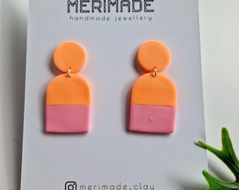 May include: A pair of handmade, two-tone earrings. Each earring features a peach-colored circle at the top, connected to a peach and pink arch shape. The earrings are displayed on a white card with the text "MERIMADE handmade jewellery" and the Instagram handle "merimade_clay".