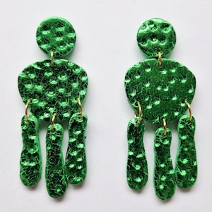 Può includere: Un paio di orecchini pendenti verde brillante. Ogni orecchino presenta una parte superiore strutturata e circolare, un pezzo centrale più grande e di forma irregolare e tre forme a goccia allungate pendenti. Gli orecchini hanno cerchi dorati e una finitura lucida e screpolata.