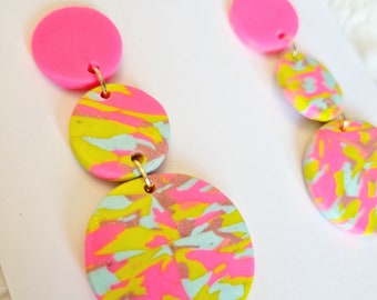 May include: A pair of pink and multi-colored dangle earrings with a marbled design. The earrings are handmade and feature a pink circle at the top, followed by two larger circles with a marbled design in pink, yellow, and blue.