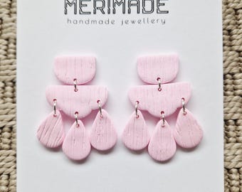 May include: A pair of light pink handmade earrings. The earrings are made of clay and feature a textured surface. The design includes a half-circle top, a curved middle section, and three teardrop-shaped dangles. The earrings are displayed on a white card with the text "MERIMADE handmade jewellery".