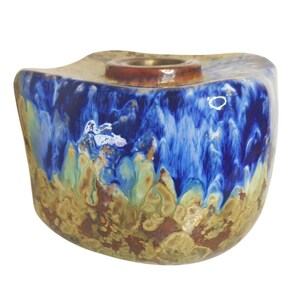 May include: A ceramic candle holder with an unusual, organic form. The holder has a mix of blue, green, and brown glaze, giving a textured, flowing look. The top has a circular opening for a candle, with a brown rim.