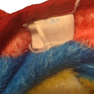 May include: Close-up of a white tag with the word "JW" printed in blue. The tag is attached to a red, blue, yellow, and pink fuzzy fabric.
