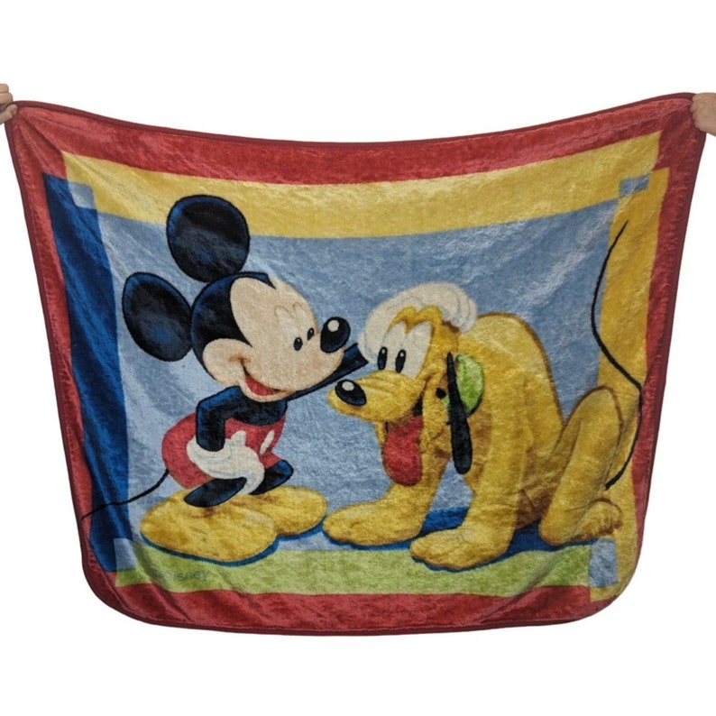 May include: A red, yellow, and blue fleece blanket with a cartoon image of Mickey Mouse and Pluto. Mickey Mouse is wearing red shorts and yellow shoes. Pluto is wearing a yellow collar and has a brown nose.