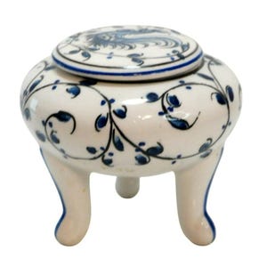May include: A small, round, cream-colored ceramic jar with a matching lid, featuring a hand-painted blue floral vine pattern. The jar is supported by three curved legs and has a smooth, glossy finish with a blue line accent on the lid edge.