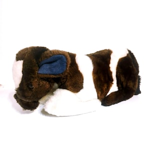 May include: A plush toy calf in a lying position. The toy features brown fur with white patches and a blue inner ear. The calf is designed to look realistic and is set against a white background.