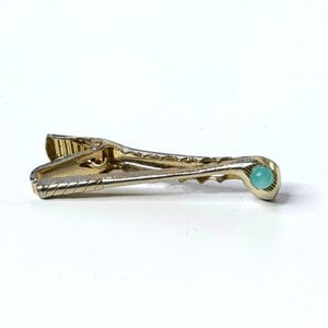 May include: A gold-toned metal tie clip shaped like a golf club. The shaft of the club features a textured, cross-hatched pattern along the handle, while the club head is accented with a small, round, light blue cabochon stone set in the centre.
