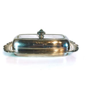 Puede incluir: Una mantequera plateada con tapa y detalles ornamentales. El plato rectangular tiene una tapa curvada con un asa decorativa. Los bordes del plato tienen un diseño festoneado.