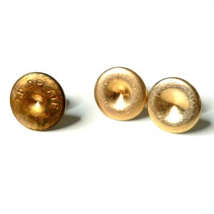 以下が含まれることがあります： 中央が凹んだ円形の金メッキボタン3つ。各ボタンの外縁には「GOLD PLATE」という浮き彫りの文字が刻まれています。ボタンは金属光沢仕上げで、1つは他の2つよりもわずかに色が濃く、変色しているように見えます。