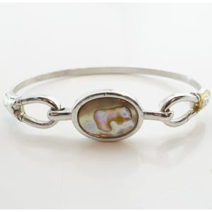 May include: Silver-tone bracelet featuring an oval mother-of-pearl centerpiece with a bear design. The bracelet has a hinged design with decorative links and gold-tone accents. The bracelet is shown on a white background.