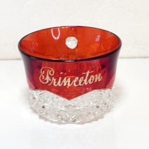Pode incluir: Uma taça de vidro vermelho profundo com a palavra Princeton gravada em letra cursiva na frente. A parte inferior da taça tem um padrão de diamante transparente e texturizado que contrasta com a secção superior vermelha translúcida.