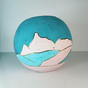 Puede incluir: Un jarrón de cerámica redondo con una parte superior turquesa texturizada y un diseño de paisaje rosa y turquesa. Líneas doradas delinean las montañas y los elementos del paisaje. Un artículo decorativo.