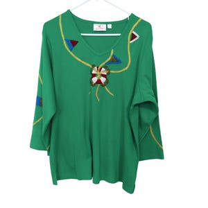 May include: Green long-sleeve shirt with a V-neck. The shirt features gold rope-like embroidery and colorful geometric shapes. A floral design is embroidered on the front. The shirt is hanging on a white hanger.