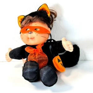 Op de afbeelding: Een pluche pop verkleed als zwarte kat met een oranje fluwelen oogmasker, bijpassende kattenoren en een doorschijnende oranje strik om de nek. De pop draagt een zwart pakje met een oranje pompoenvormig tasje aan de zijkant bevestigd.