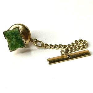 May include: A gold-tone tie tack featuring a square-shaped green stone inlay with a mottled texture. The tie tack is connected by a short gold-tone chain to a horizontal bar clasp, designed to secure the accessory to a garment or necktie.
