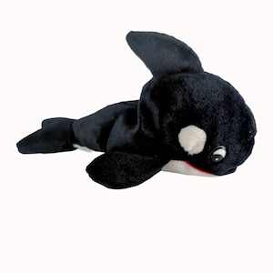 May include: A plush toy orca whale in black and white. The orca has a white patch near its eye and a red mouth. The toy is lying down on a white background. The orca has a dorsal fin and flippers.