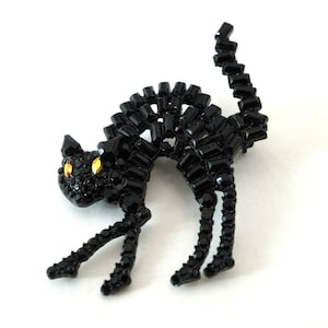 Puede incluir: Broche de gato negro con la espalda curvada y la cola levantada. El gato está hecho de piedras negras rectangulares y redondas, con ojos amarillos. El broche está sobre un fondo blanco.