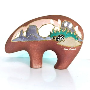 May include: A brown ceramic bear figurine featuring a Southwestern landscape design. The design includes purple mountains, a white sky, and green and gold accents. The artist's signature, Vera Russell, is visible.