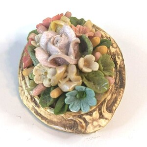 Puede incluir: Un broche floral vintage con una rosa rosa y otras flores en tonos de amarillo, blanco y verde. El broche está engastado en un marco de metal dorado con un diseño texturizado.