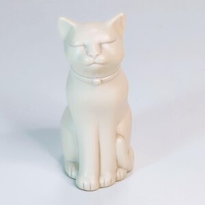 May include: A cream-colored cat figurine in a seated position with closed eyes. The ceramic cat wears a collar with a small decorative element. The sculpture has a smooth finish and is set against a white background.