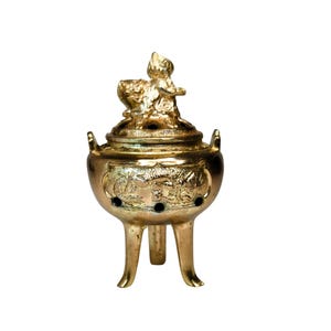 May include: A gold-colored incense burner with a three-legged base and two handles. The burner features intricate dragon and floral designs, with a detailed figure on the lid. The burner has three small holes on the front.