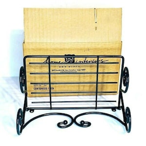 May include: Black metal magazine rack with a scroll design. The rack is sitting on a brown cardboard box with the text "Home Interiors and Gifts" printed on it.