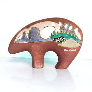 May include: A ceramic bear figurine in shades of brown, with a landscape design. The design features purple mesas, green foliage, and a white sky, outlined in gold. The artist's signature, Vera Russell, is visible on the side.