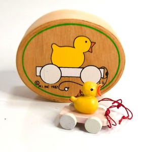 May include: A vintage wooden toy set featuring a yellow duck. The set includes a round wooden box with a duck illustration and a pull-along toy duck on a pink cart with white wheels. The box has a green border and the text "M.J. INC 1982".