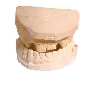 May include: A beige dental mold of a human mouth, showcasing both upper and lower teeth. The model is a detailed replica, with visible teeth and gum lines. The surface has a slightly rough texture, and some text is visible on the top portion.