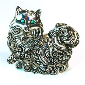 Puede incluir: Figura de gato plateado con intrincados adornos y ojos verdes brillantes. El gato está representado en posición sentada, con la cola enroscada. La figura es de metal brillante y tiene una superficie texturizada con patrones intrincados.