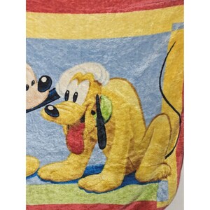 May include: A yellow and brown plush blanket with a cartoon image of Pluto the dog. Pluto is sitting on a blue background with a red, yellow, and green border.