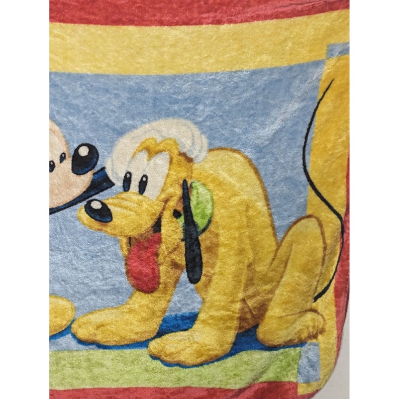 May include: A yellow and brown plush blanket with a cartoon image of Pluto the dog. Pluto is sitting on a blue background with a red, yellow, and green border.