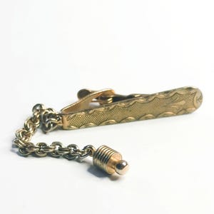 May include: A gold-toned tie clip with an engraved design and a chain attached to a decorative end piece. The clip has a textured pattern and a clasp. The end piece has a cylindrical shape with a small ball at the end.