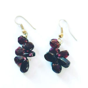 May include: A pair of dangle earrings featuring clusters of dark red, polished stones. The stones are attached to gold-colored ear wires. The earrings are set against a white background.