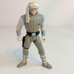 Op de afbeelding: Vintage actiefiguur van een personage in een lichtgrijze en witte sneeuwoverall met een zwarte riem en holster. De figuur heeft een helm met gele bril en staat met gespreide benen.