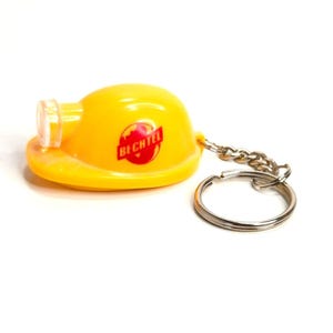 Puede incluir: Un llavero de casco amarillo con un llavero y una cadena plateados. El casco tiene un logotipo rojo y blanco que dice "BECHTEL". El llavero está sobre un fondo blanco.