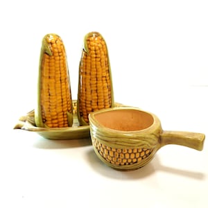 May include: A ceramic set featuring two corn-shaped salt and pepper shakers standing on a matching corn-shaped tray, accompanied by a small, handle-equipped corn-textured bowl with a light tan interior and a glossy green and yellow finish.